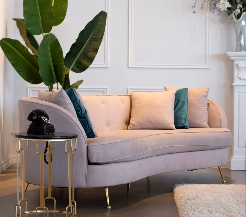 Curved beige sofa with green and beige cushions, gold metal side table with black vintage telephone, green plant in background in an elegant living room.