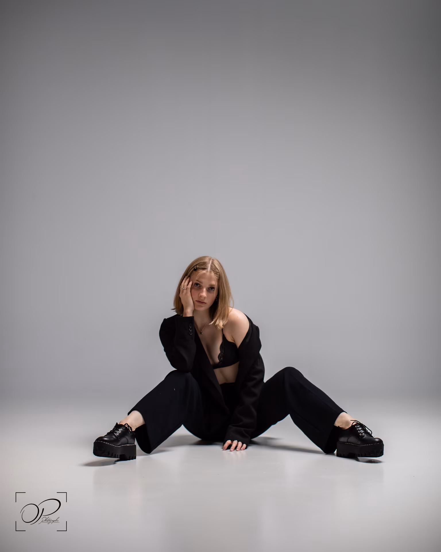 Young woman sitting on the floor, legs apart, wearing an open black blazer over a black bra and thick-soled black shoes.